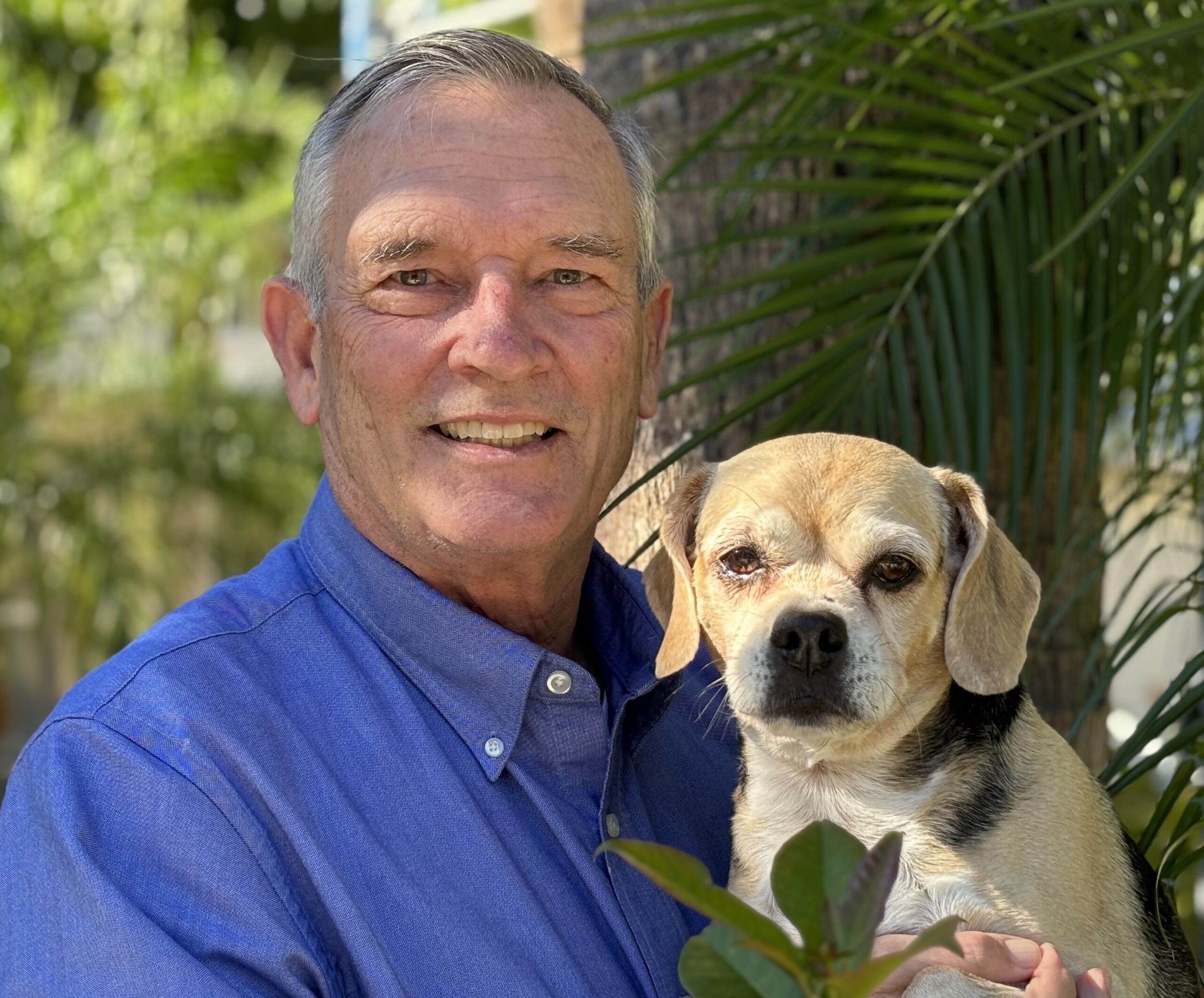 GH Ellis in a blue shirt smiling and holding a small dog outdoors, with green foliage in the background."