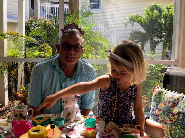 GH Ellis wearing a blue shirt and a crown, sitting at a tea party with his young granddaughter who is reaching for food. They are outdoors with lush greenery in the background.