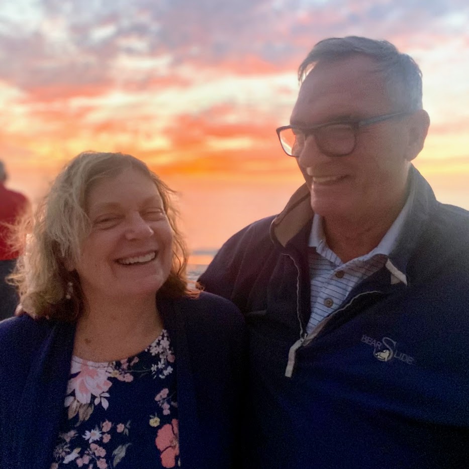 GH Ellis and his wife Jan smiling at each other on the beach at sunset in Naples, Florida. The sky is filled with vibrant colors.