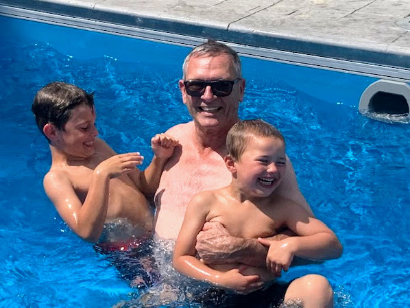 GH Ellis wearing sunglasses, swimming in a pool with his two grandsons. They are all smiling and having fun.